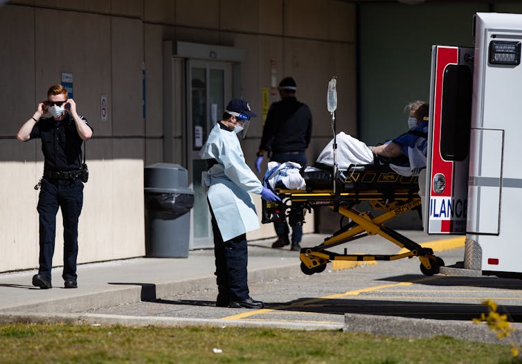 El estándar revisado de respiradores de CSA debería ayudarnos a respirar mejor 1 Un paramédico que lleva una mascarilla saca una camilla de una ambulancia mientras otro paramédico se pone una mascarilla en el fondo.