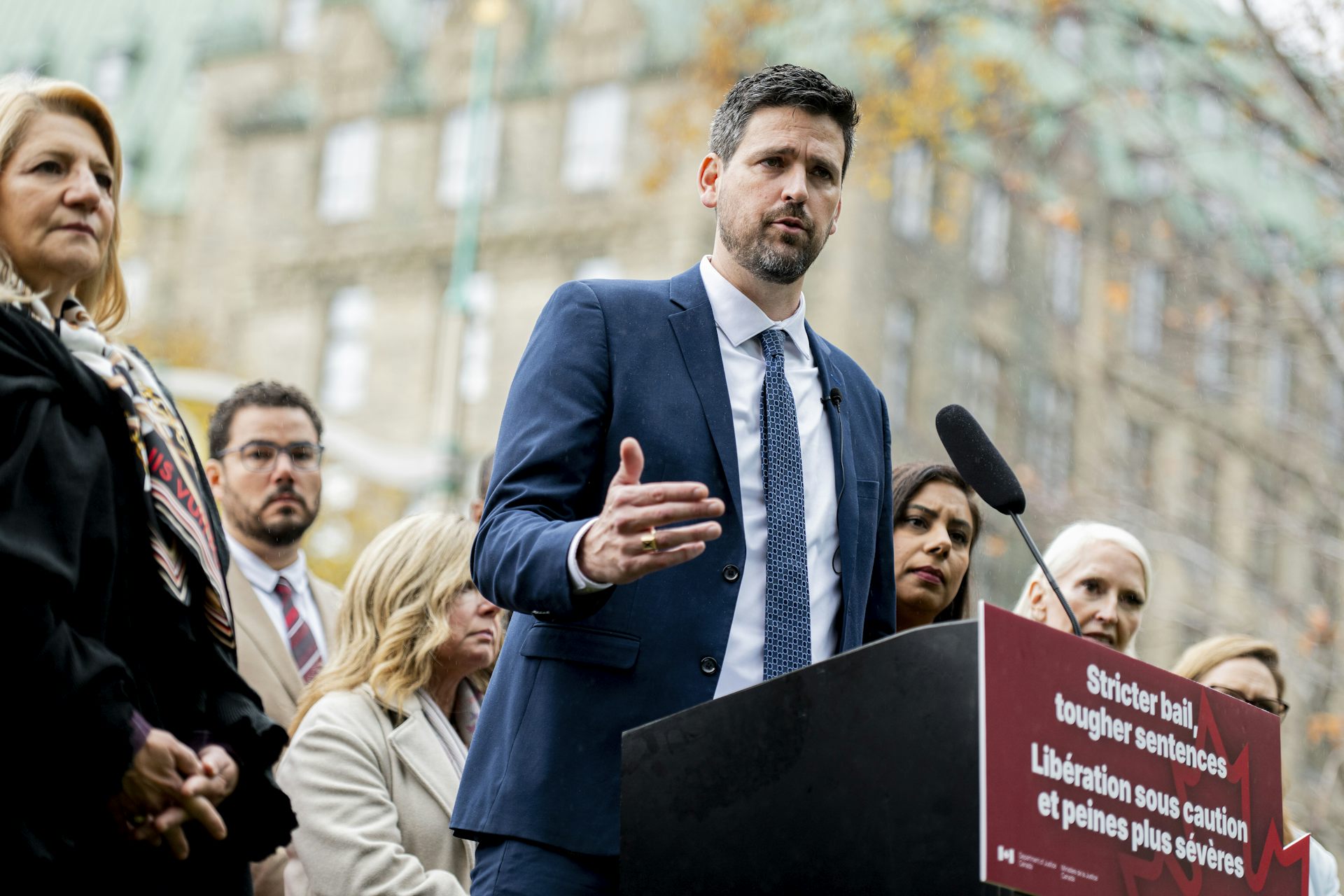 A tall man with dark hair and beard speaks from behind a lectern that says stricter bail, tougher sentences in English and French.