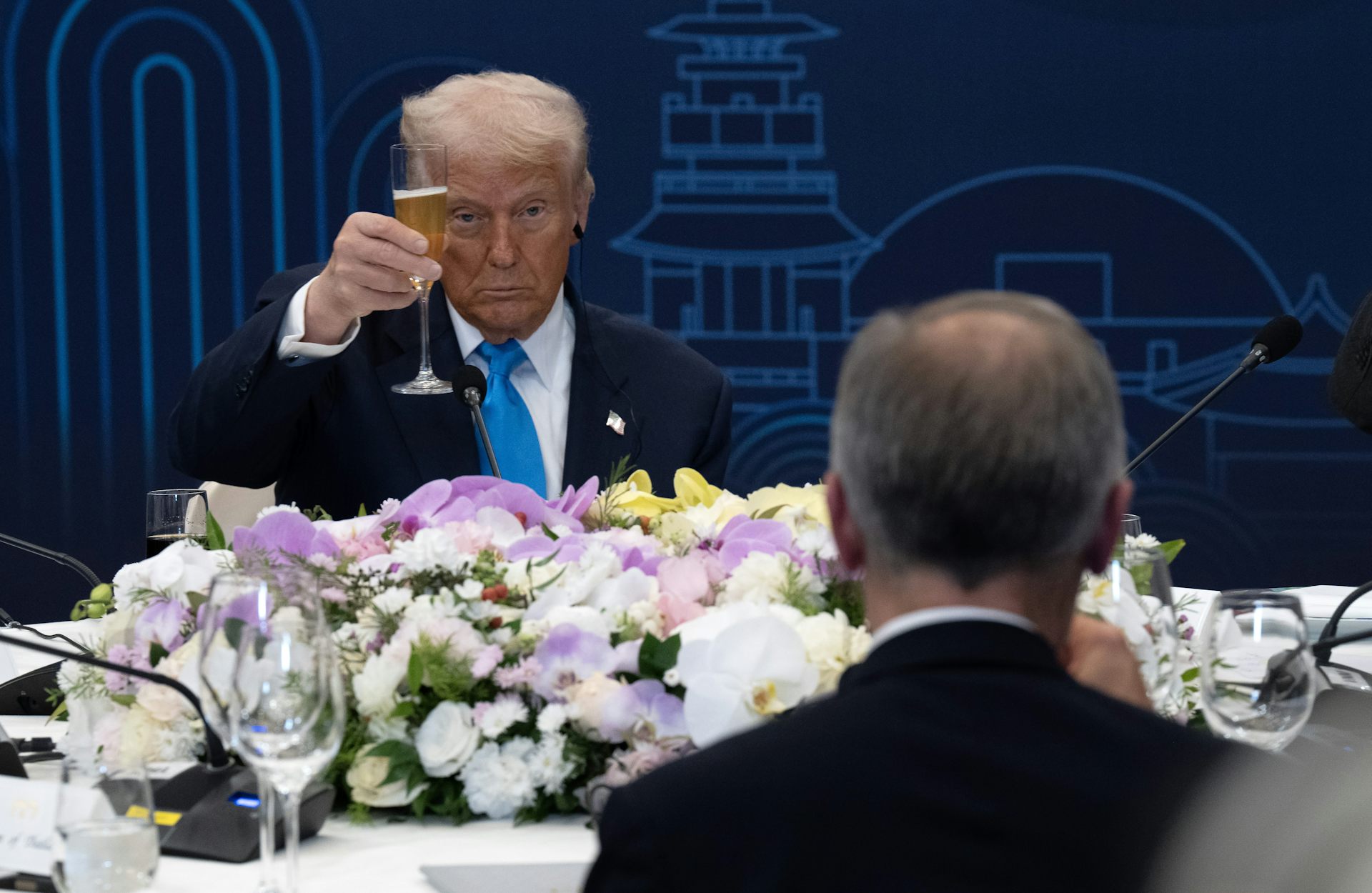 A man with fluffy yellow-ish white hair raises his champagne glass at another man whose back is to the camera.