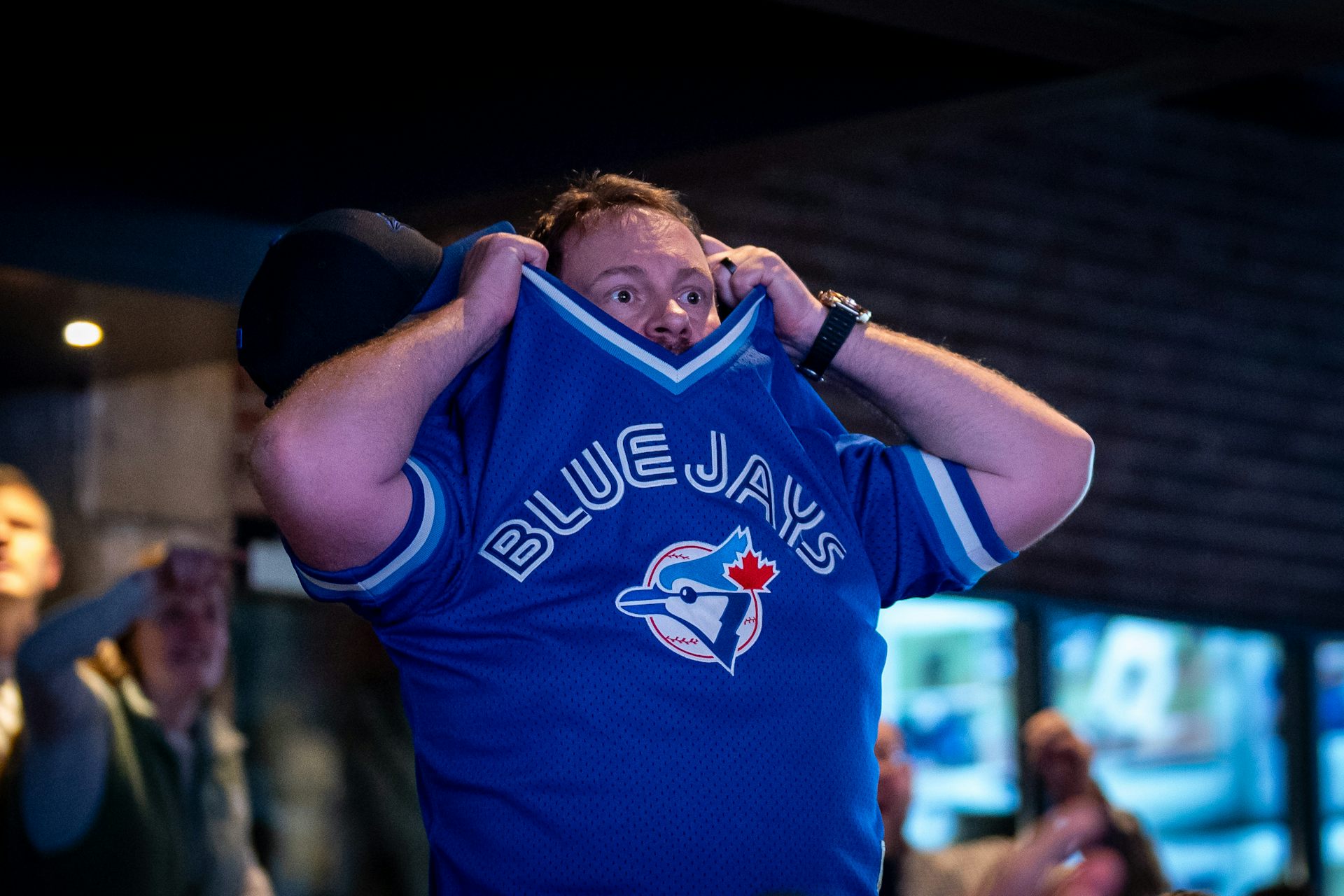 A man holds his Blue Jays jersey almost over his head as he stares at the screen in despair.