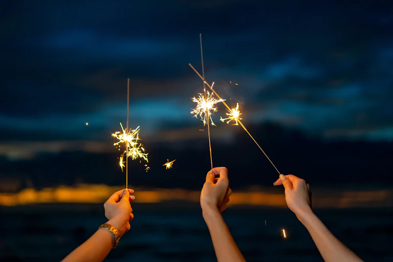 Three hands holding lit sparklers on a dark night.