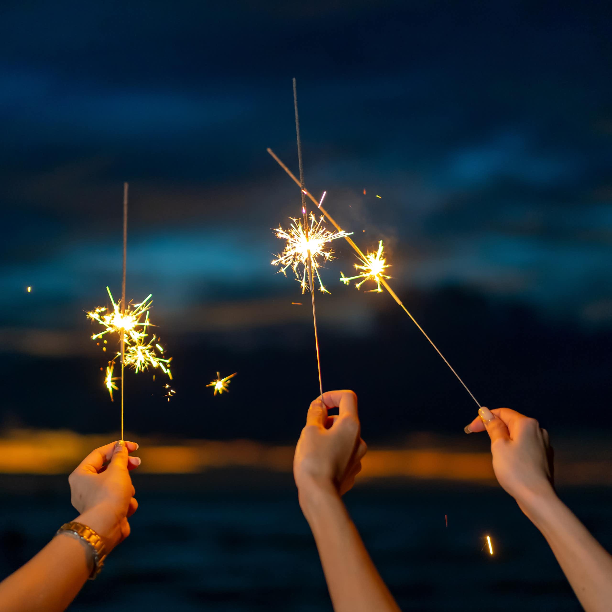 Three hands holding lit sparklers on a dark night.