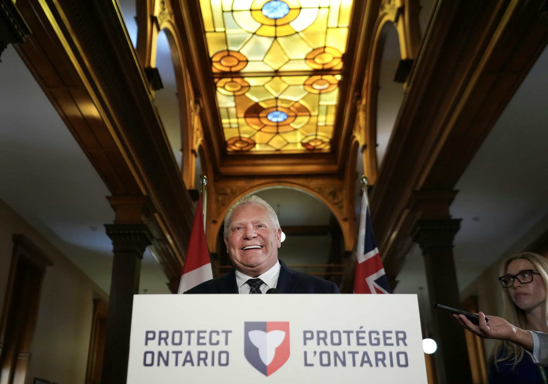 Un hombre sonriente y corpulento con cabello gris y ralo se encuentra detrás de un atril que dice Proteger Ontario en francés e inglés. Sobre su cabeza hay una vidriera dorada.