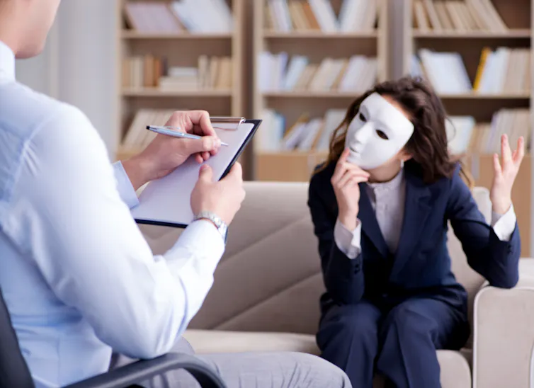 Woman in suit sits on a sofa opposite a therapist taking notes. The woman is wearing a white mask.