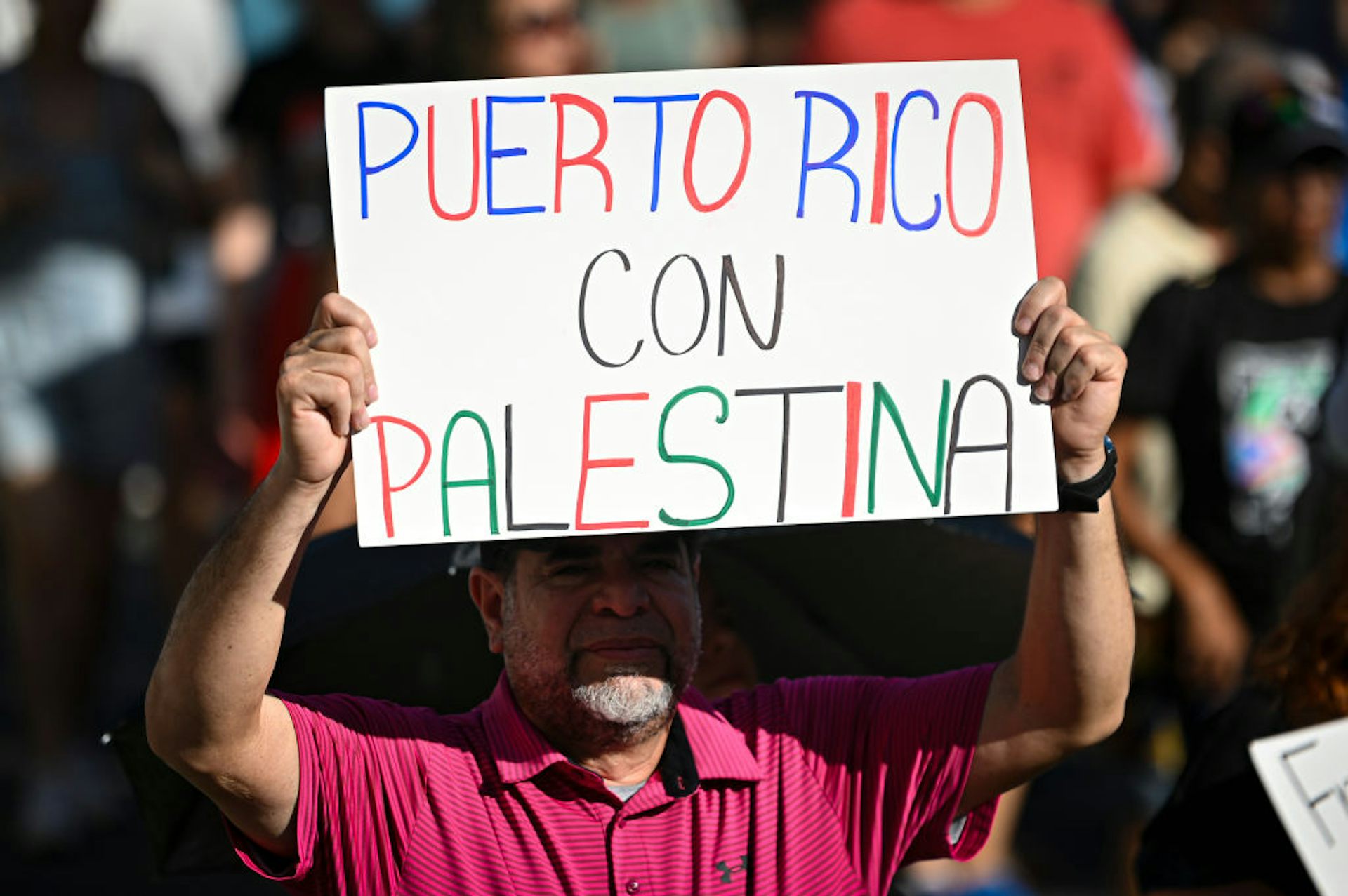 A man in a red shirt, standing amid a crowd, holds a sign that says, in Spanish, 'Puerto Rico with Palestine.'