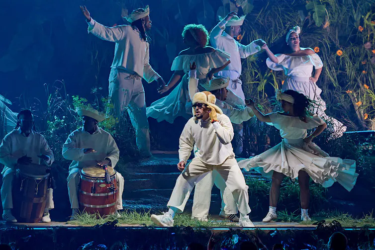 A man in a white costume holds his hand up to his ear, looking off stage, as dancers in white outfits move behind him and beat drums.