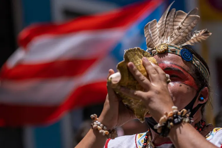 A man in a feather headdress blows into a large shell, standing in front of a Puerto Rican flag.