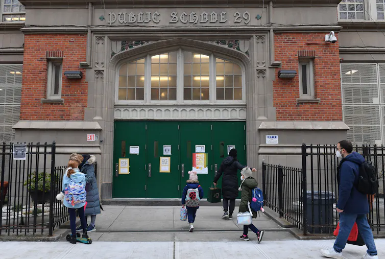 Several children and adults walk into a large brick building with green doors.