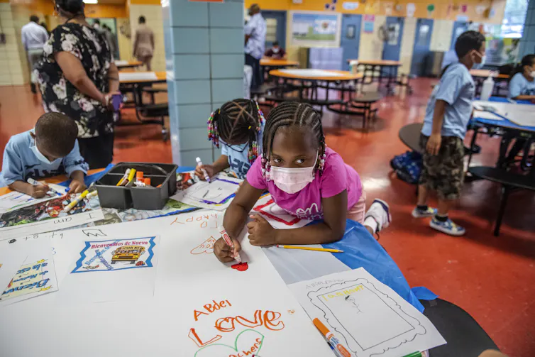 A group of young children wearing face masks sit at a table and color on white paper.
