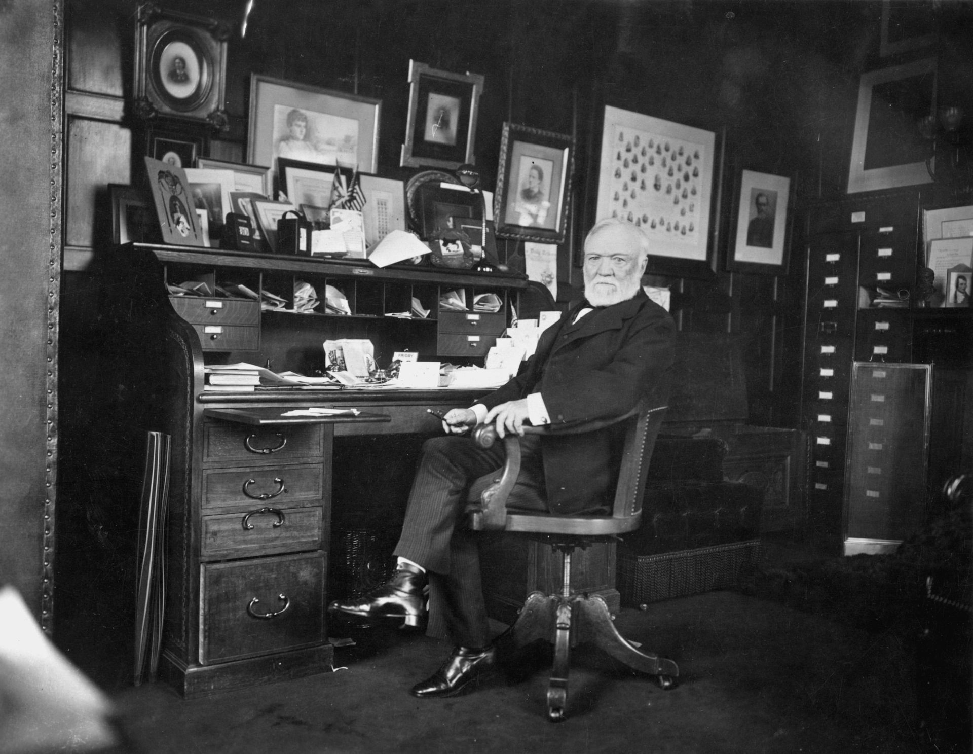 Andrew Carnegie sits at his writing desk in front of lots of family photos.