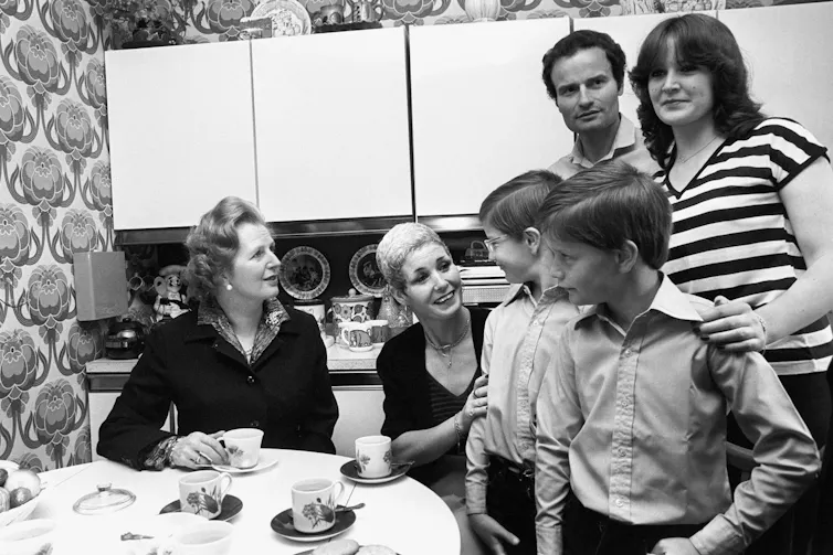 Prime minister Margaret Thatcher (left) drinks tea with a family in their council house kitchen.