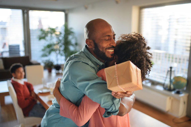A bald man with a beard smiles widely as he hugs a girl with curly hair, while he holds a wrapped package in one hand.