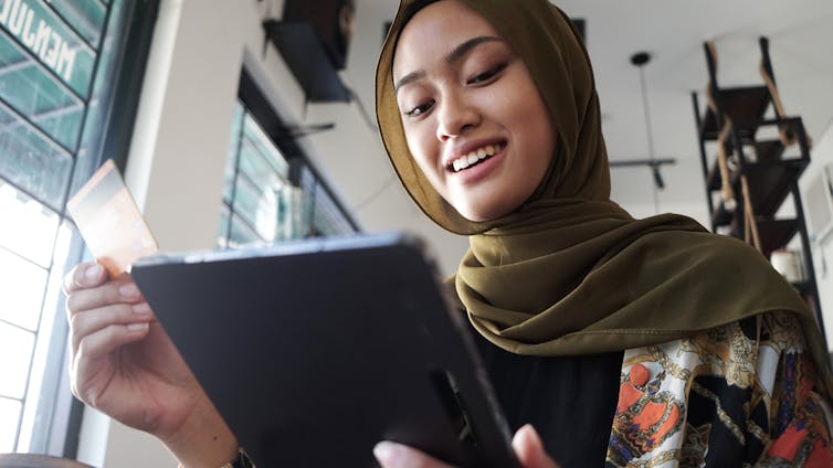 'A woman wearing a green headscarf and holding a credit card in one hand smiles as she looks down at a tablet.