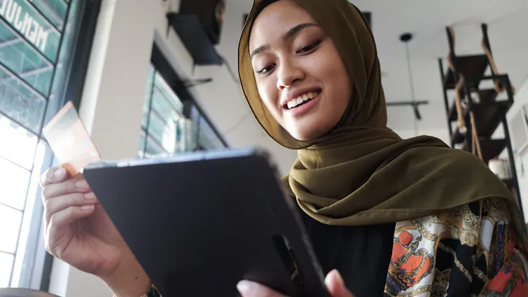 'A woman wearing a green headscarf and holding a credit card in one hand smiles as she looks down at a tablet.