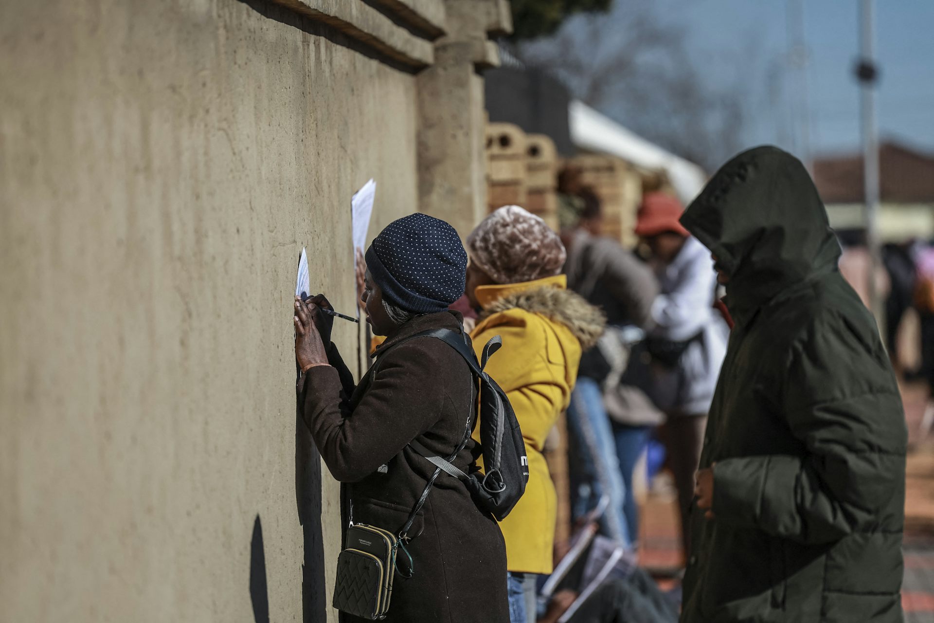 Women write on pieces of paper, pressing against a wall