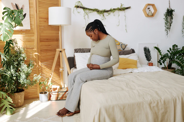 A pregnant woman sits on her bed looking pensive, her hand resting on her stomach.