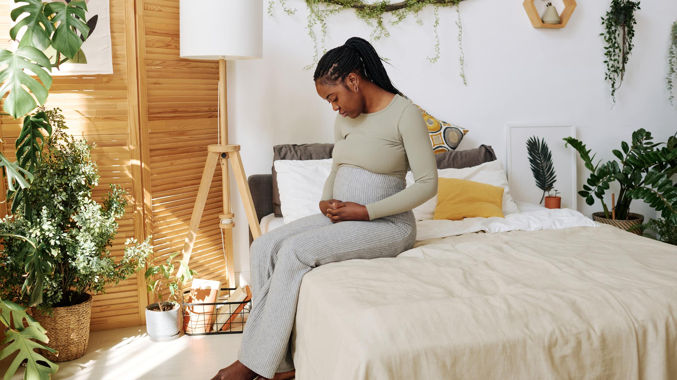 A pregnant woman sits on her bed looking pensive, her hand resting on her stomach.