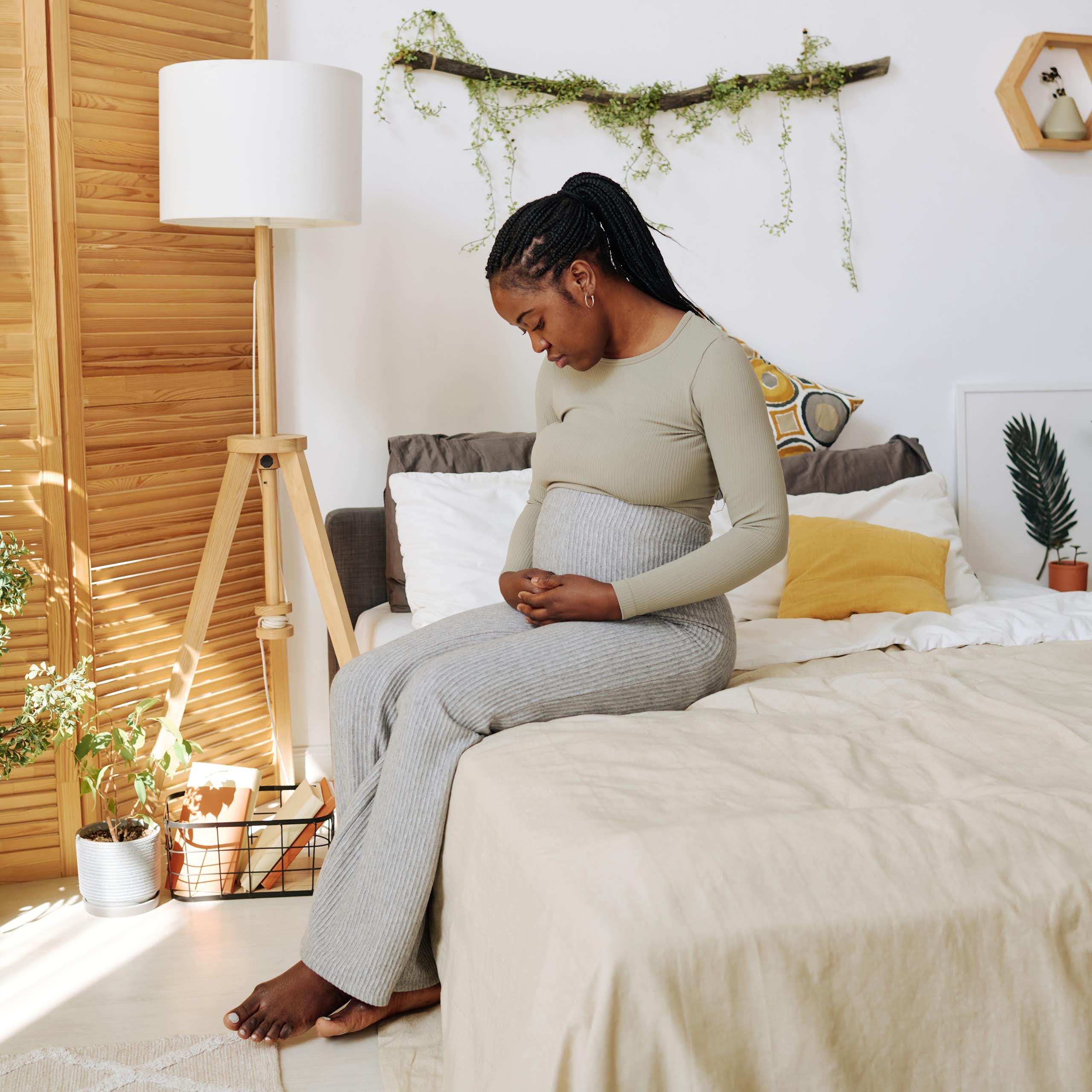 A pregnant woman sits on her bed looking pensive, her hand resting on her stomach.