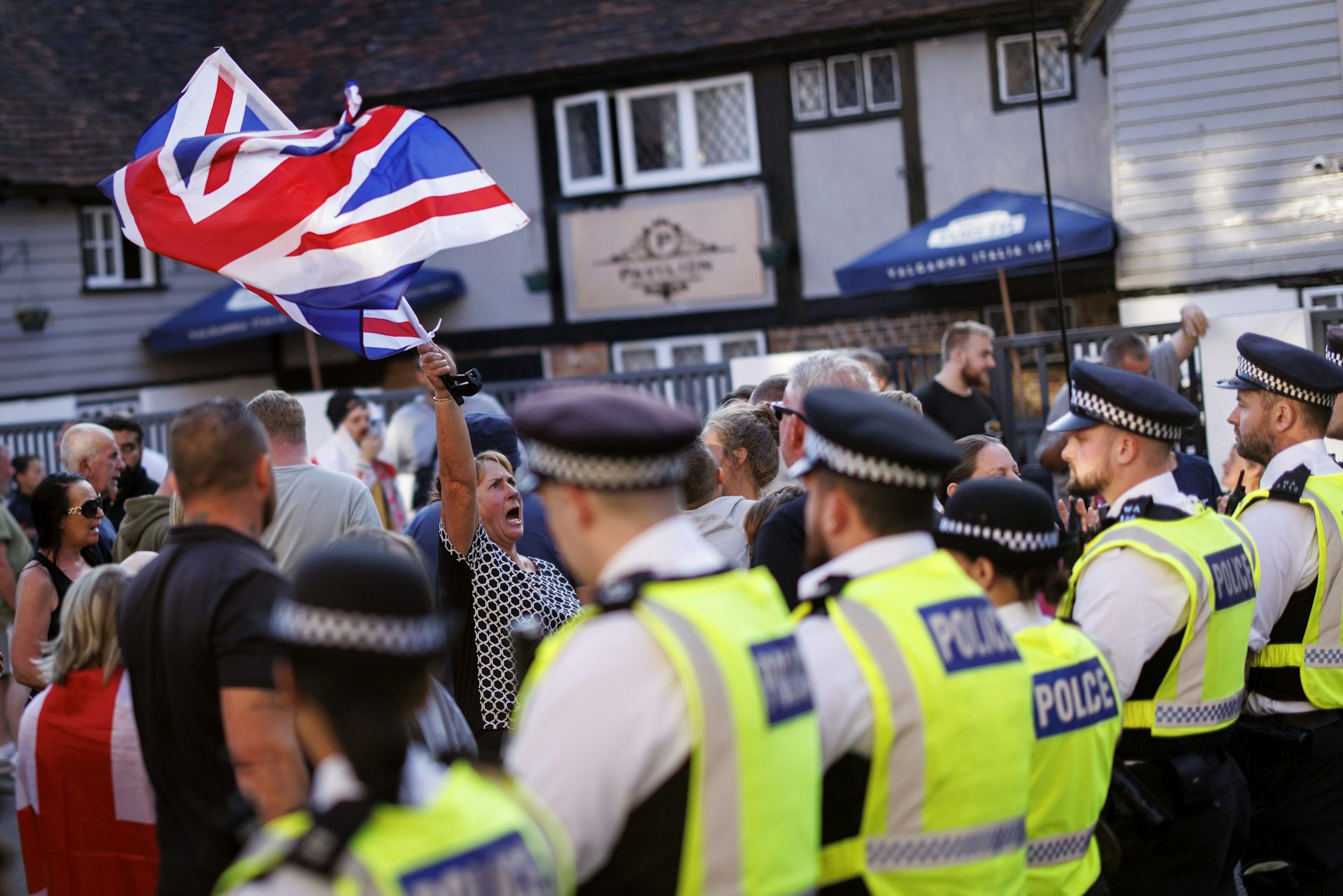 Police and protesters waving a large union jack flag outside of a hotel housing asylum seekers