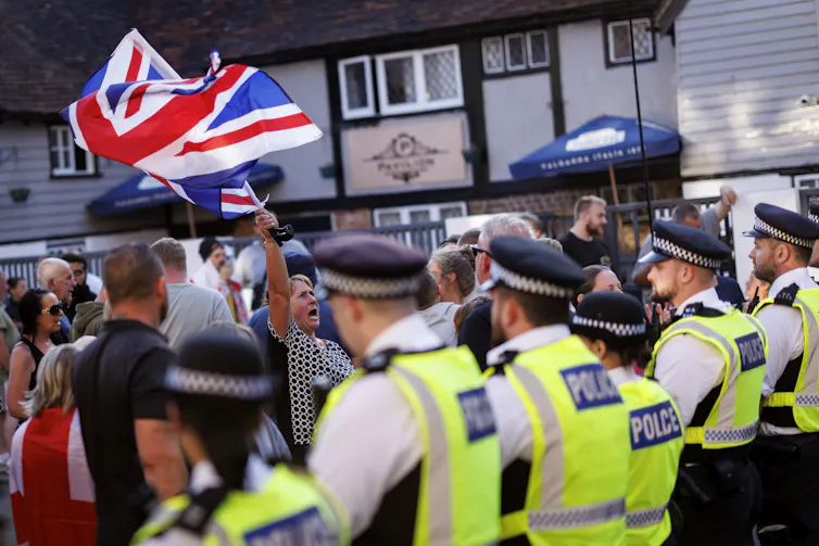 Police and protesters waving a large union jack flag outside of a hotel housing asylum seekers