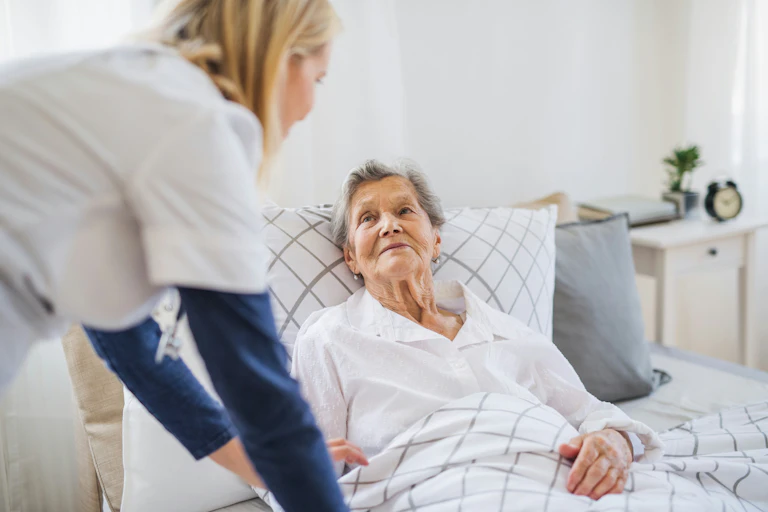 A senior woman in bed with a care person by her side.