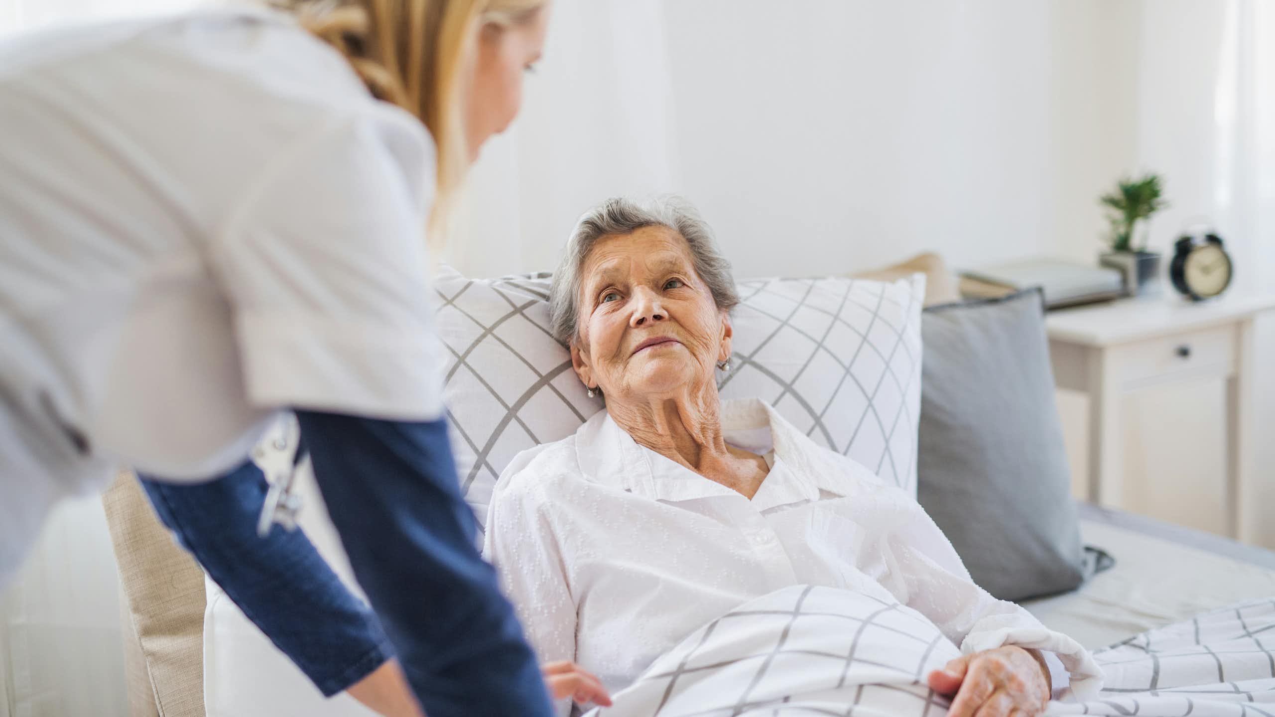 A senior woman in bed with a care person by her side.