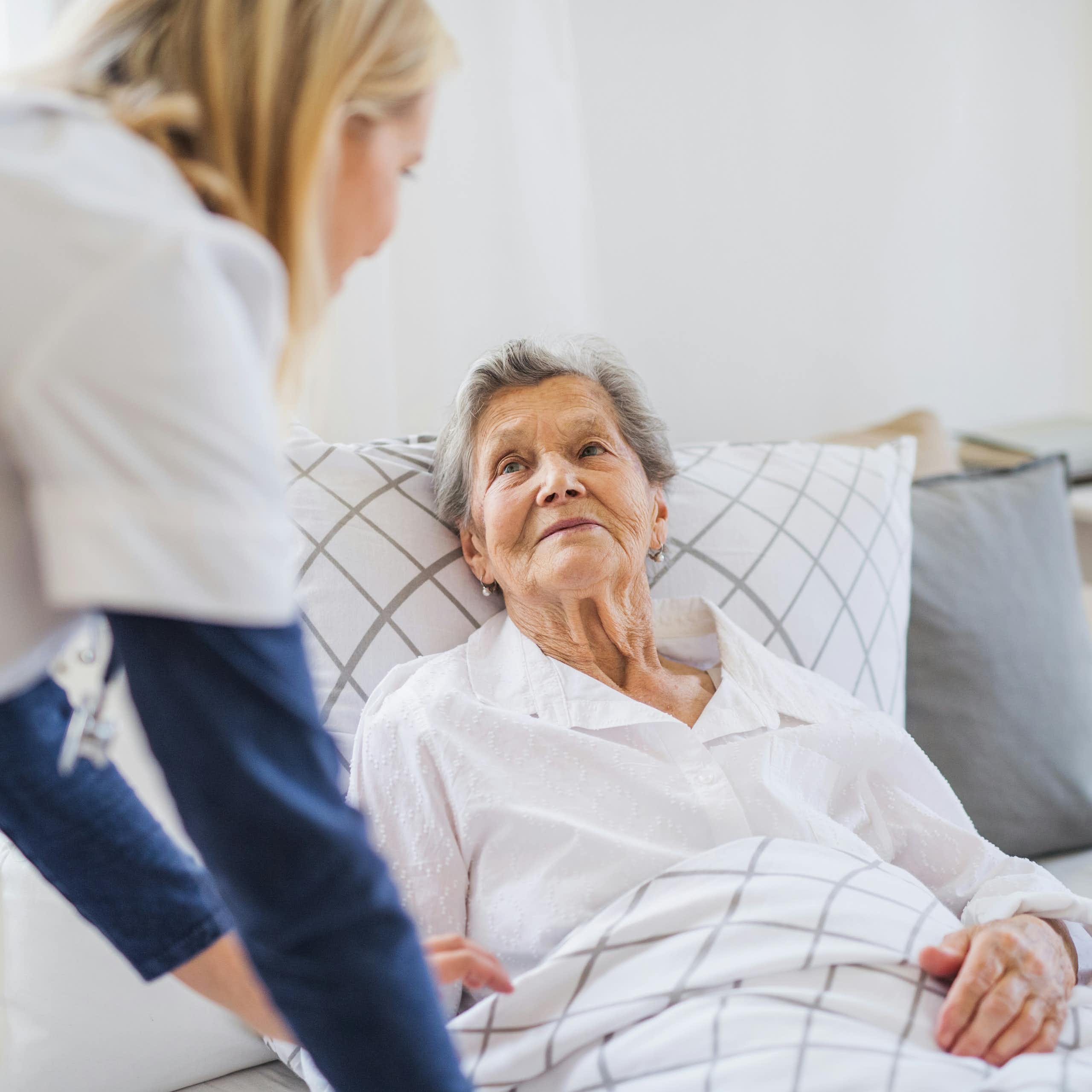 A senior woman in bed with a care person by her side.