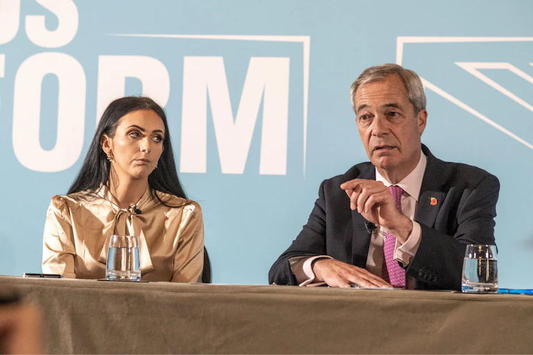Reynolds and Farage sit side by side behind a table, on stage in front of a Reform UK wall