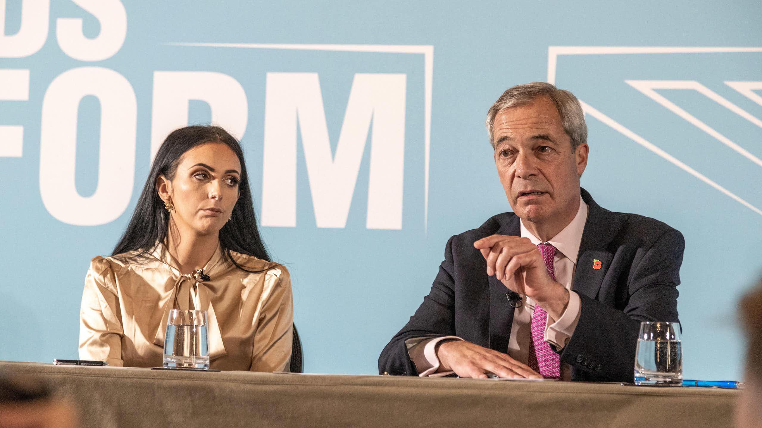Reynolds and Farage sit side by side behind a table, on stage in front of a Reform UK wall