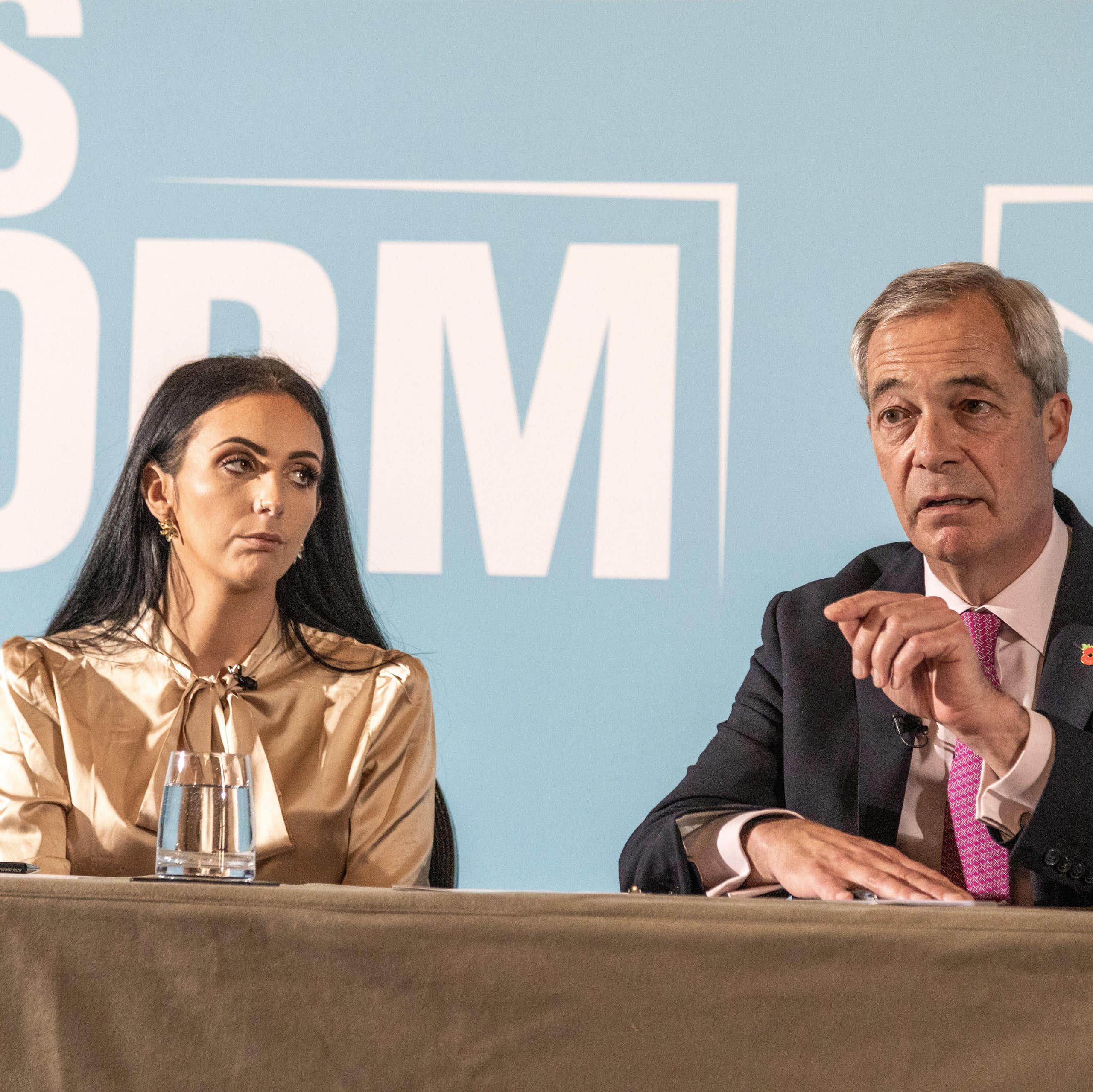 Reynolds and Farage sit side by side behind a table, on stage in front of a Reform UK wall