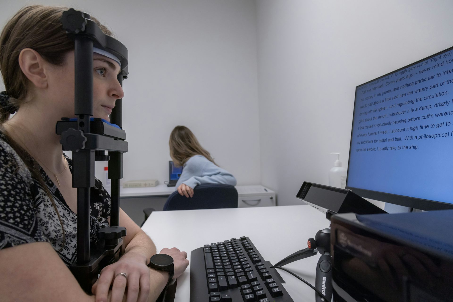 A woman reads text on a screen while placing her chin on a headrest, with a small camera in front of her below the screen.
