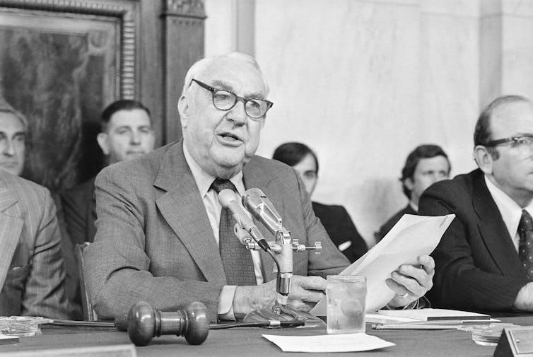 A white-haired man, wearing glasses and holding a sheet of paper, sits at a dais speaking into a microphone.
