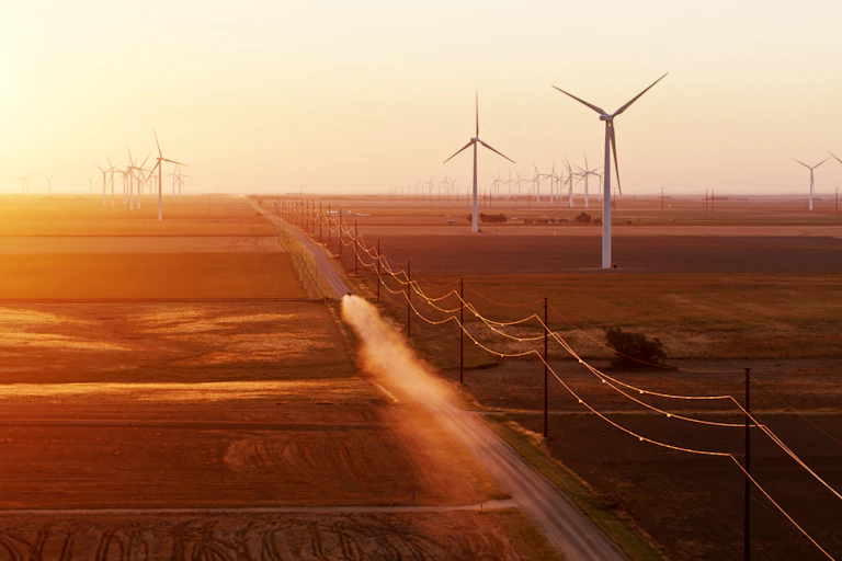 A truck kicks up dust on a dirt road running through farm fields with wind turbines.