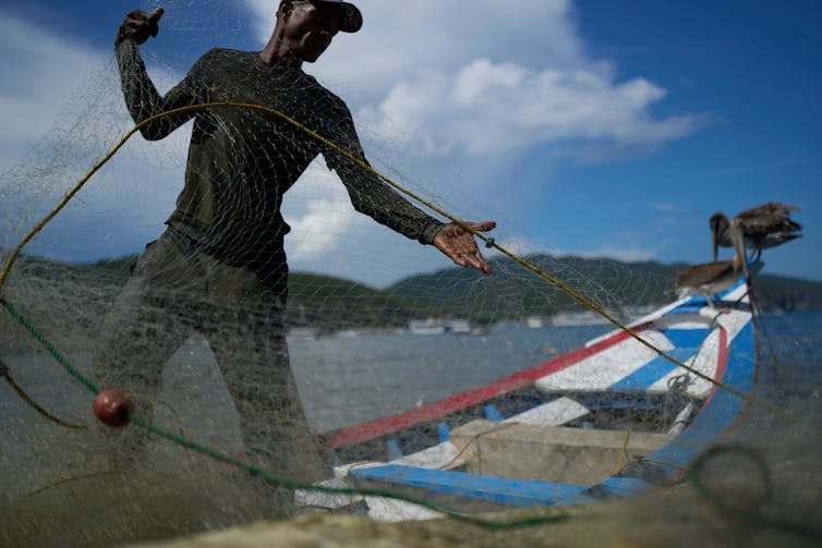 Un pescador lleva una red y se encuentra junto a un barco cerca del agua.
