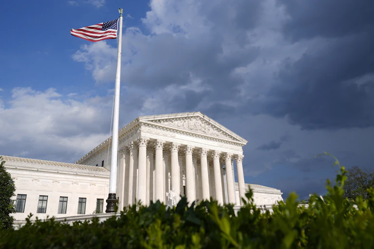 The Supreme Court building, photographed from a ways away, with a blue but cloudy sky and an American flag in front of the building.