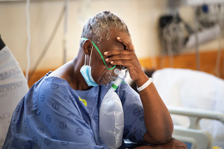 A woman in a hospital gown, wearing an oxygen mask, holding her head in her hand