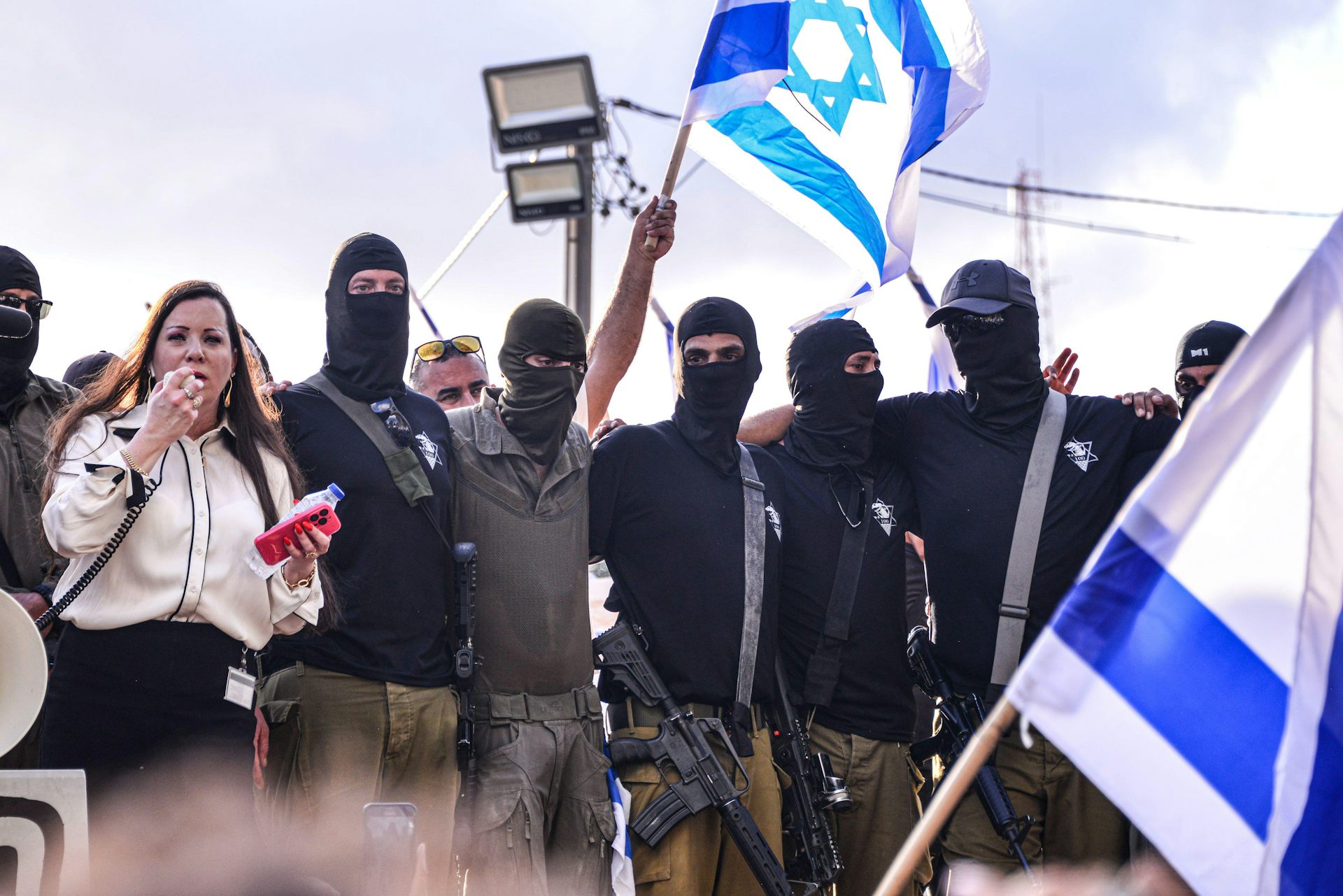 An Israeli woman speaks into a loudhailer and is flanked by men in balaclavas holding Israeli flags.