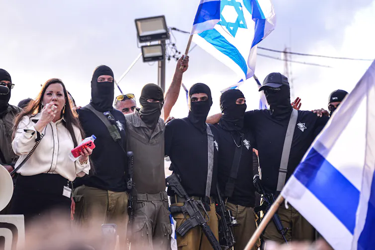 An Israeli woman speaks into a loudhailer and is flanked by men in balaclavas holding Israeli flags.