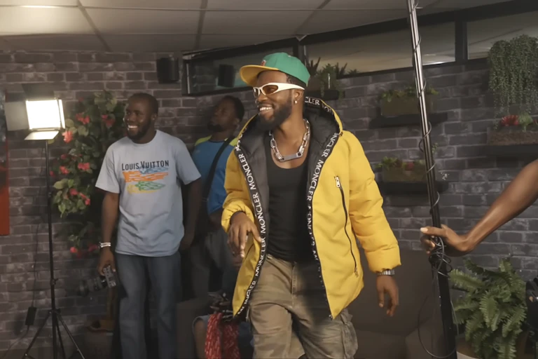 A bearded African man smailes in a cap and yellow jacket in what looks like a studio with other men, smiling, one holding recording equipment.
