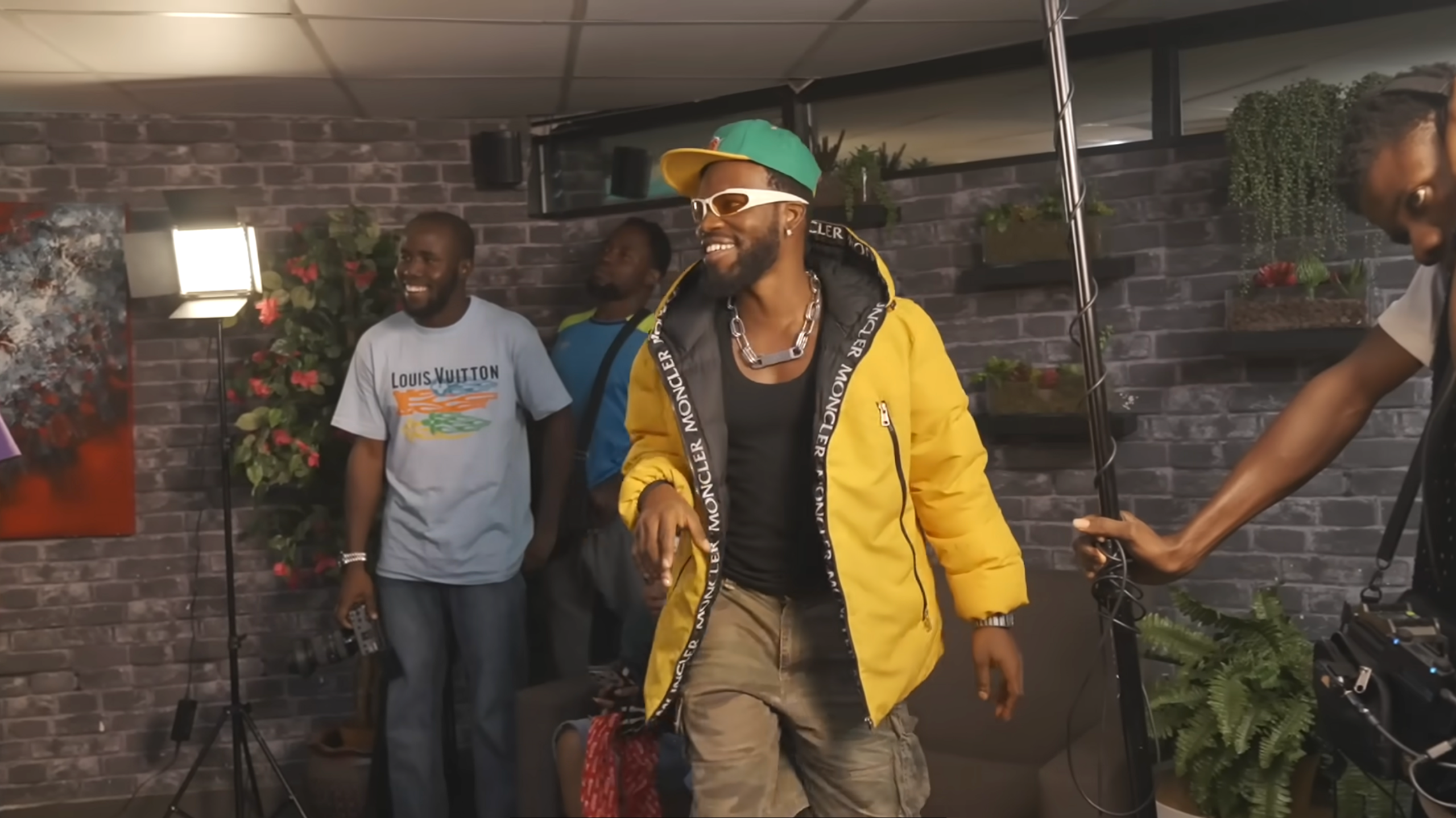 A bearded African man smailes in a cap and yellow jacket in what looks like a studio with other men, smiling, one holding recording equipment.