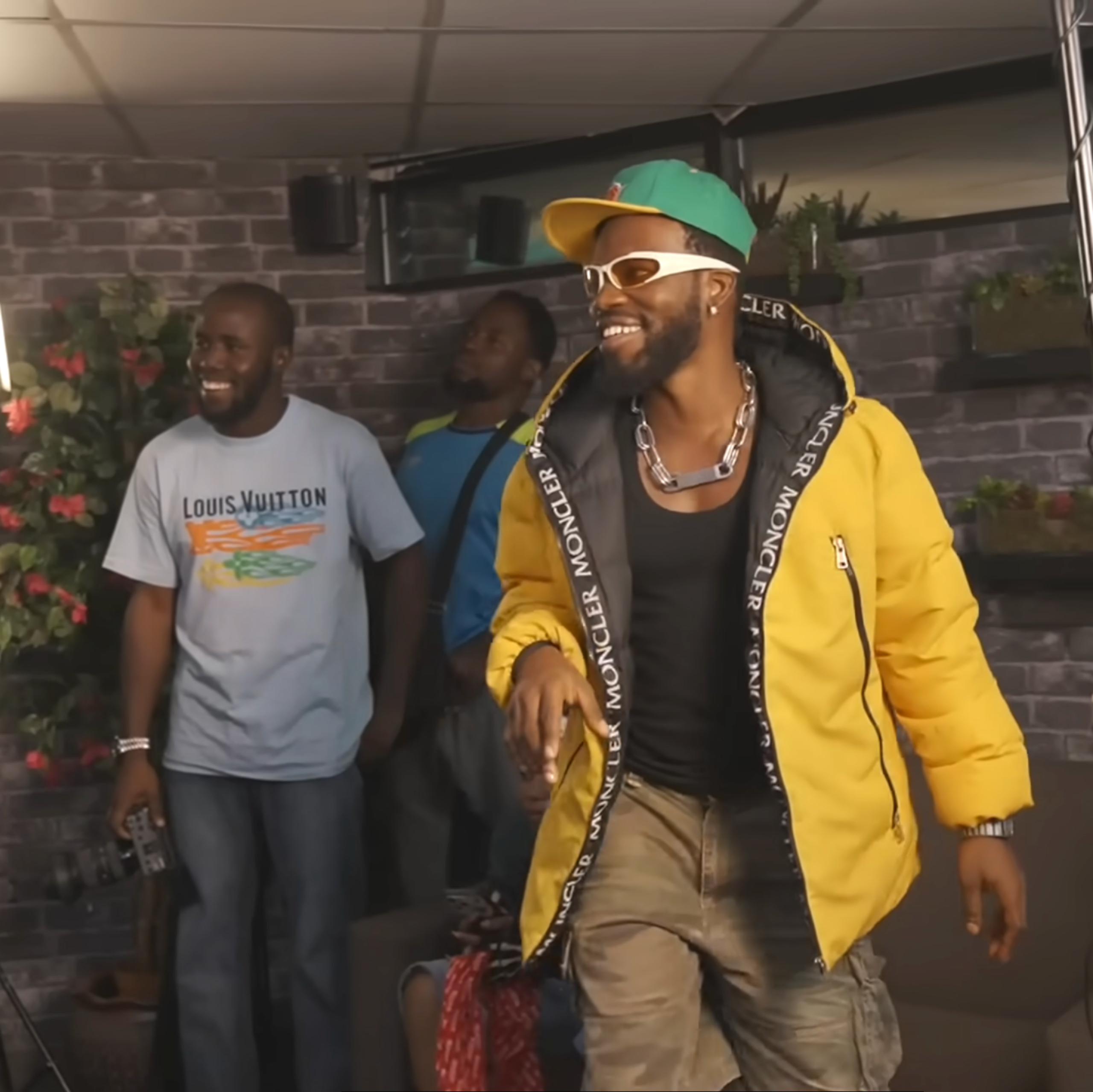 A bearded African man smailes in a cap and yellow jacket in what looks like a studio with other men, smiling, one holding recording equipment.