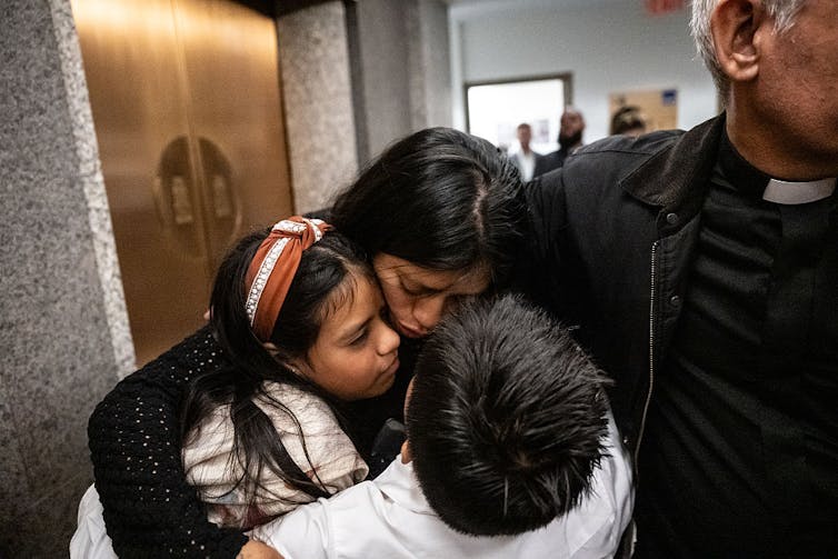 Un hombre con alzacuello de sacerdote, cuyo rostro está fuera de cuadro, rodea con su brazo a una mujer angustiada y a dos niños vestidos de blanco.