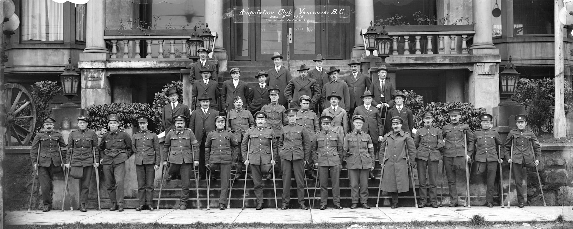 Men in overcoats in rows on steps, holding single crutches, many with an amputated leg. 
