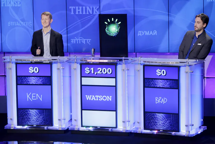 Black screen with IBM Watson logo on a Jeopardy stand with $1,200 stood between two contestants with $0 each