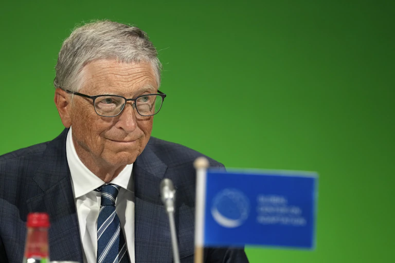 An elderly man wearing glasses and a dark suit sitting in front of a microphone