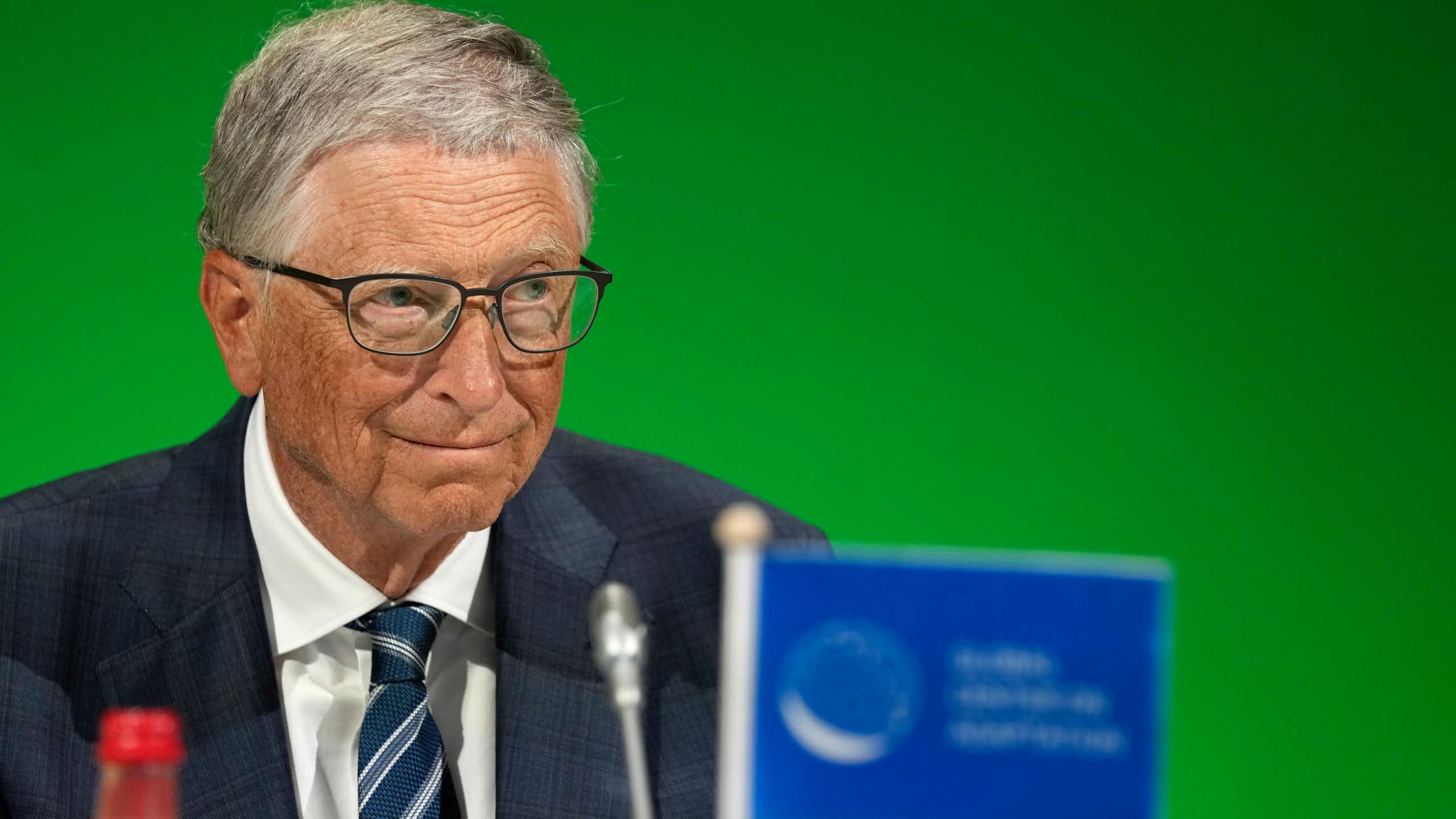 An elderly man wearing glasses and a dark suit sitting in front of a microphone