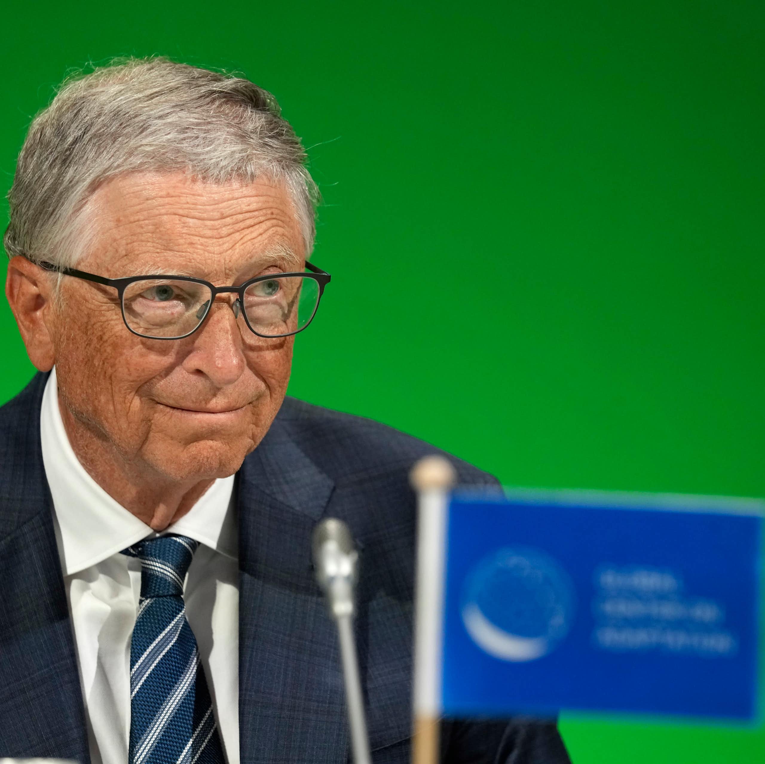 An elderly man wearing glasses and a dark suit sitting in front of a microphone