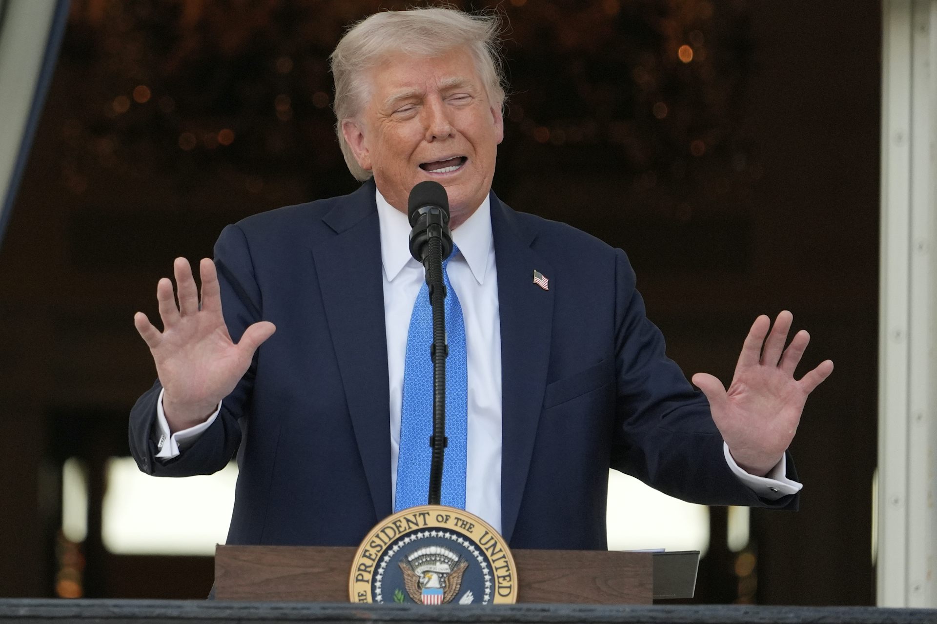 An old orange-hued man in a dark suit giving a speech while raising his arms