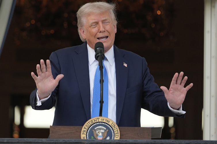 An old orange-hued man in a dark suit giving a speech while raising his arms