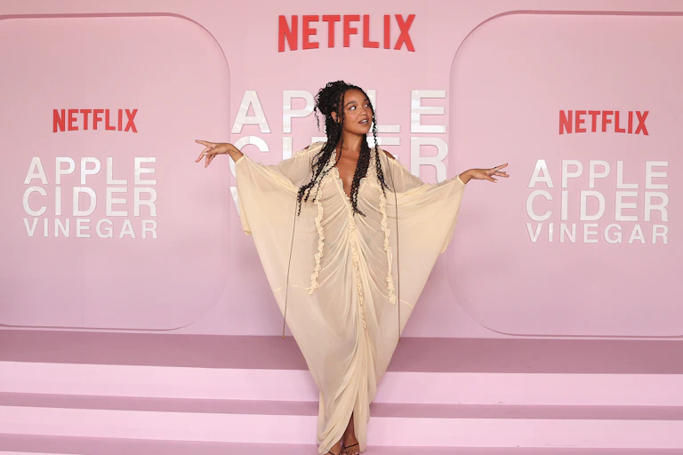 Actress Aisha Dee stands in front of a promotional pink backdrop for the Netflix series Apple Cider Vinegar.